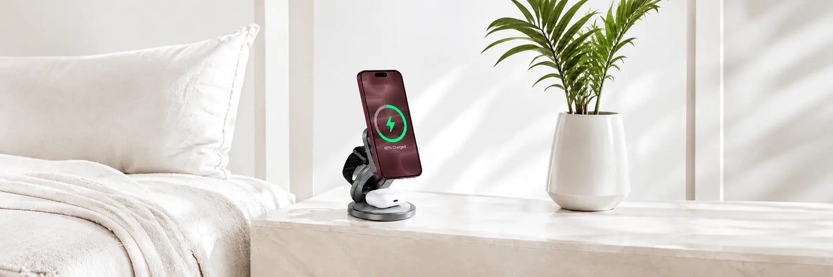 Foldable wireless charging station with a burgundy phone, watch and earbuds beside a bed.