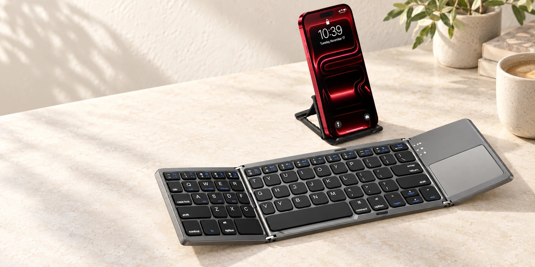 Gray folding wireless keyboard open beside a phone on a small stand.